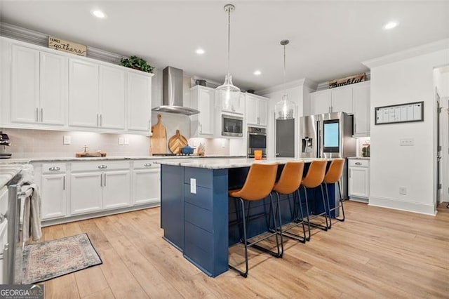 kitchen with pendant lighting, wall chimney range hood, light hardwood / wood-style floors, appliances with stainless steel finishes, and a kitchen island