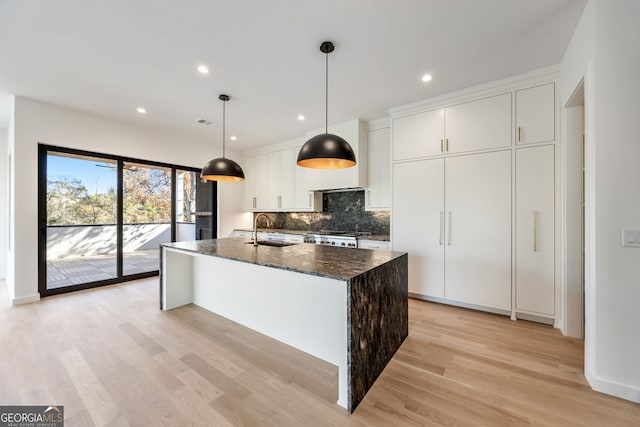 kitchen featuring dark stone counters, decorative light fixtures, white cabinets, light hardwood / wood-style floors, and an island with sink