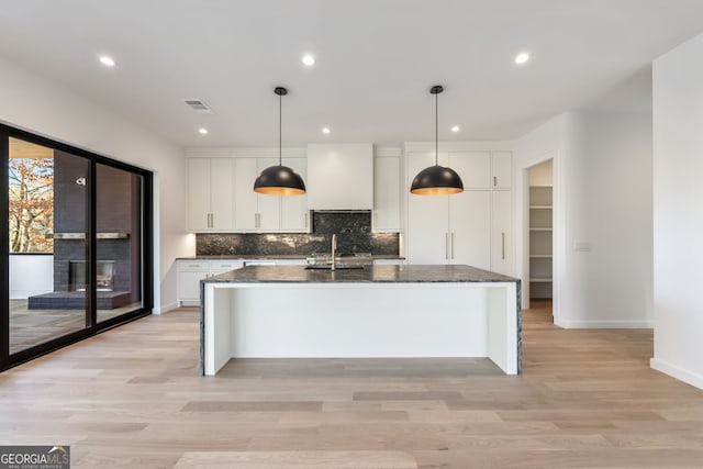 kitchen with dark stone counters, pendant lighting, a kitchen island with sink, white cabinets, and custom exhaust hood