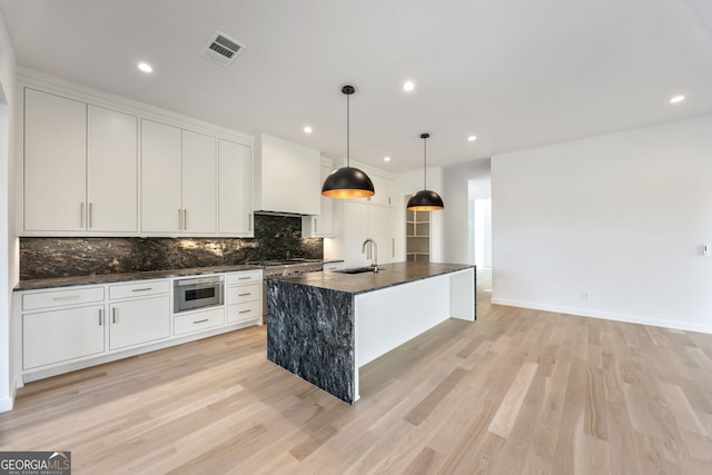 kitchen featuring light wood-type flooring, sink, decorative light fixtures, white cabinetry, and an island with sink