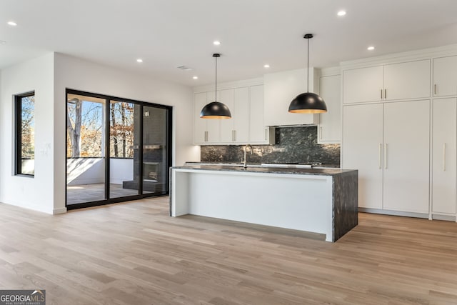 kitchen with pendant lighting, backsplash, a kitchen island with sink, white cabinets, and light hardwood / wood-style flooring