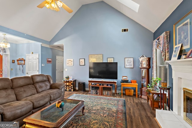 living room featuring a tile fireplace, a skylight, high vaulted ceiling, wood-type flooring, and ceiling fan with notable chandelier