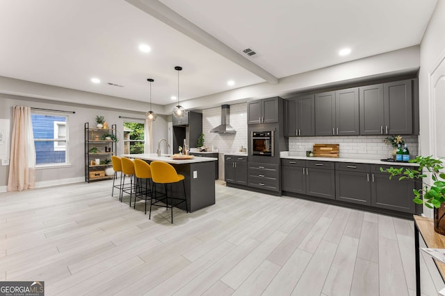 kitchen featuring a kitchen breakfast bar, stainless steel oven, wall chimney range hood, pendant lighting, and an island with sink