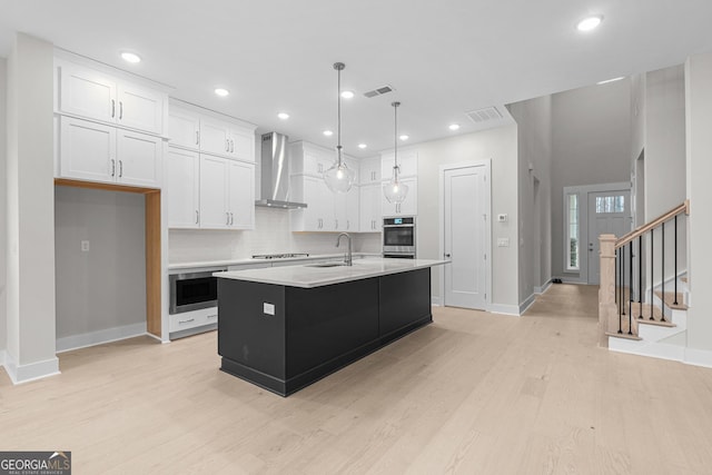 kitchen featuring wall chimney range hood, light hardwood / wood-style flooring, white cabinetry, an island with sink, and decorative light fixtures