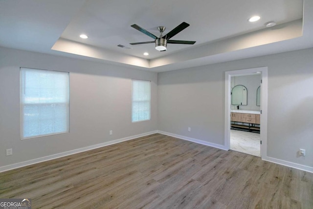 unfurnished bedroom featuring a raised ceiling, ensuite bath, ceiling fan, and light wood-type flooring