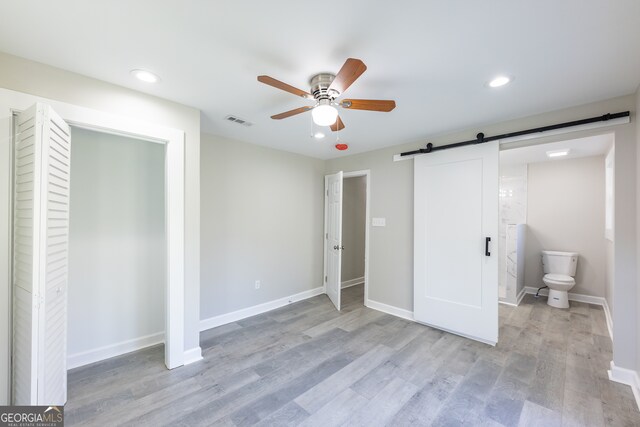 unfurnished bedroom with ceiling fan, a barn door, connected bathroom, and light hardwood / wood-style flooring