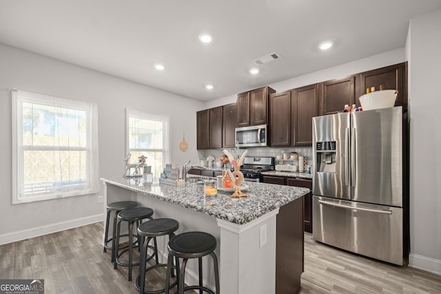 kitchen featuring appliances with stainless steel finishes, an island with sink, a kitchen bar, light stone counters, and light hardwood / wood-style flooring