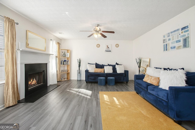 living area featuring dark wood-style floors, ceiling fan, a textured ceiling, a lit fireplace, and baseboards