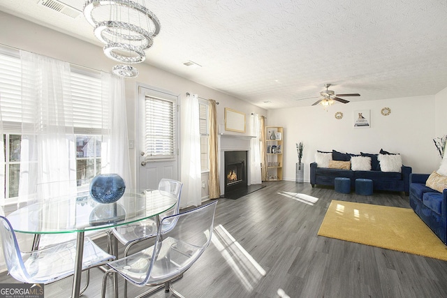 dining area with a warm lit fireplace, visible vents, dark wood-style flooring, a textured ceiling, and ceiling fan with notable chandelier