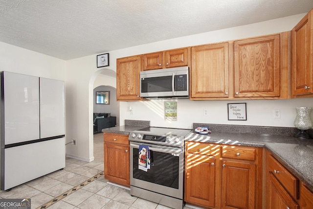 kitchen with arched walkways, stainless steel appliances, dark countertops, and brown cabinets