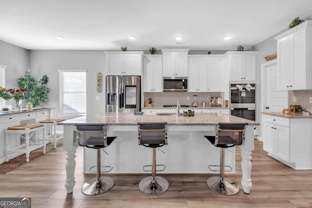 kitchen with light wood-style flooring, an island with sink, white cabinets, light stone countertops, and appliances with stainless steel finishes