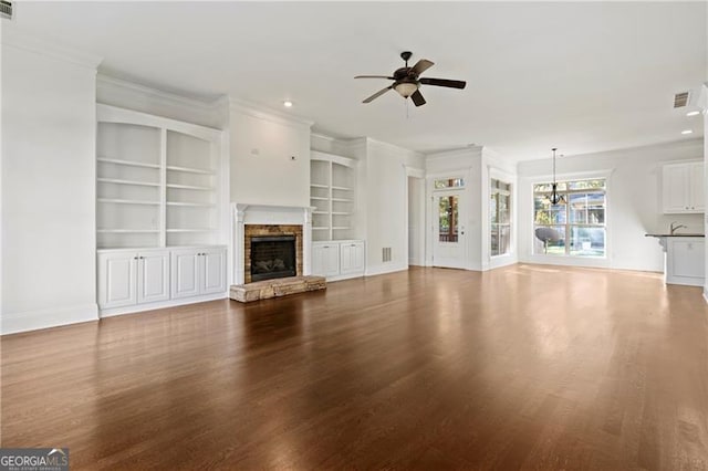 unfurnished living room featuring ornamental molding, built in shelves, ceiling fan with notable chandelier, hardwood / wood-style floors, and a stone fireplace