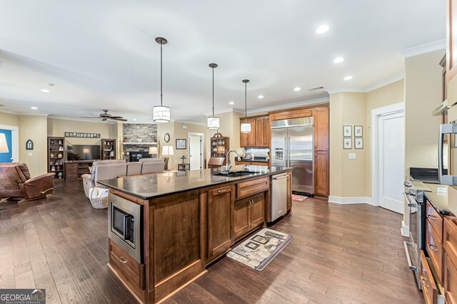 kitchen with dark wood-type flooring, sink, hanging light fixtures, built in appliances, and an island with sink