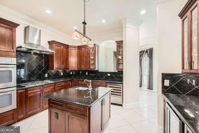 kitchen with stainless steel gas stovetop, a center island with sink, wall chimney range hood, double oven, and beverage cooler