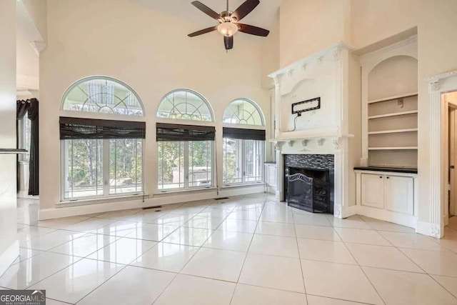 unfurnished living room with ceiling fan, built in features, light tile patterned floors, a towering ceiling, and a tiled fireplace