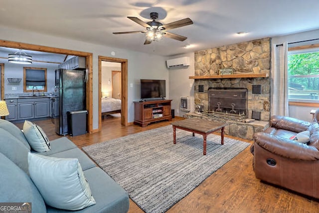 living room featuring ceiling fan, sink, a stone fireplace, a wall mounted AC, and wood-type flooring