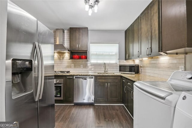 kitchen with wall chimney exhaust hood, appliances with stainless steel finishes, light stone countertops, washing machine and dryer, and a sink