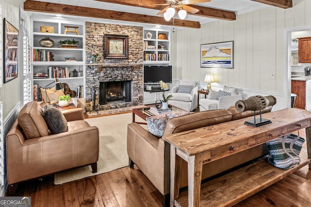 living room featuring a stone fireplace, ceiling fan, wood-type flooring, built in shelves, and beamed ceiling