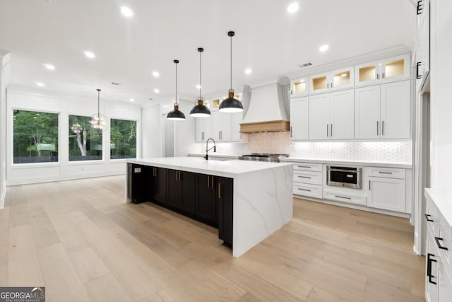 kitchen featuring a large island with sink, decorative light fixtures, custom range hood, and light hardwood / wood-style floors