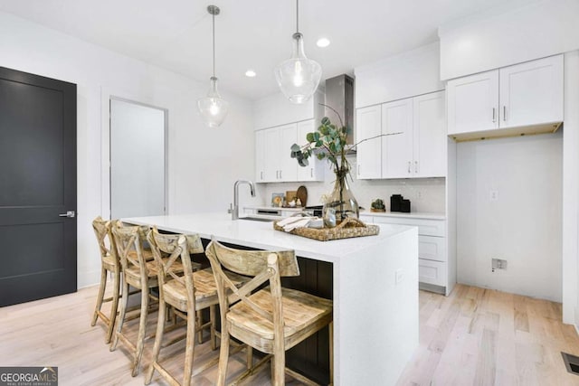 kitchen featuring wall chimney exhaust hood, a kitchen island with sink, pendant lighting, white cabinets, and light hardwood / wood-style floors