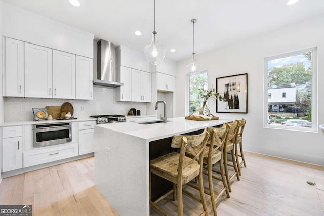 kitchen featuring stainless steel appliances, wall chimney range hood, decorative light fixtures, white cabinets, and an island with sink