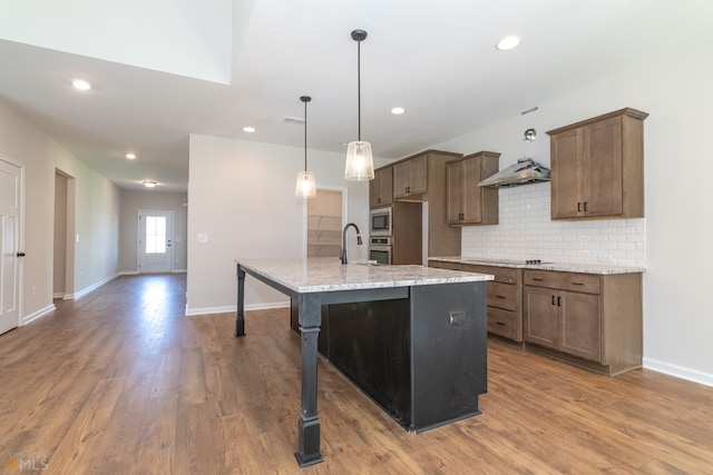 kitchen with light stone countertops, stainless steel appliances, an island with sink, light hardwood / wood-style floors, and decorative light fixtures