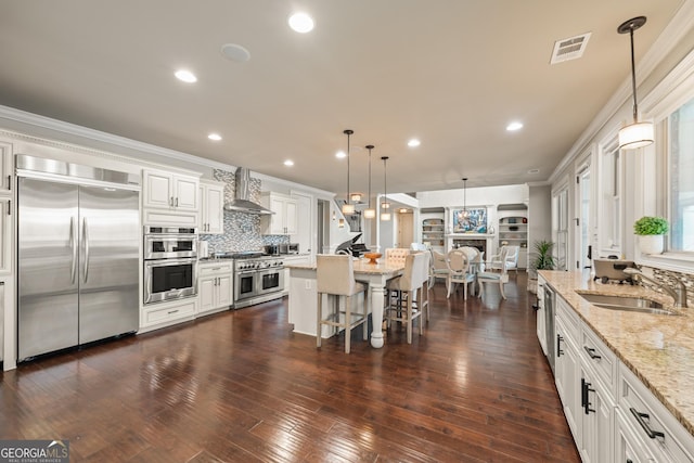 kitchen with light stone countertops, appliances with stainless steel finishes, white cabinets, wall chimney exhaust hood, and pendant lighting