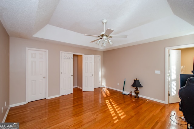 unfurnished bedroom featuring multiple closets, ceiling fan, wood-type flooring, a textured ceiling, and a tray ceiling