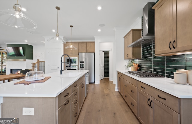 kitchen featuring sink, stainless steel appliances, wall chimney range hood, a kitchen island with sink, and light wood-type flooring