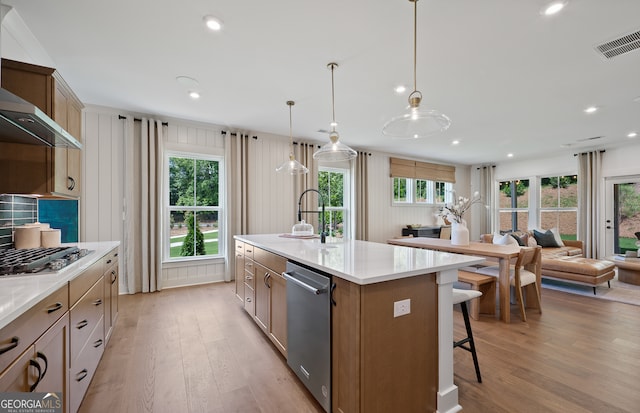 kitchen with wall chimney range hood, an island with sink, light wood-type flooring, decorative light fixtures, and appliances with stainless steel finishes