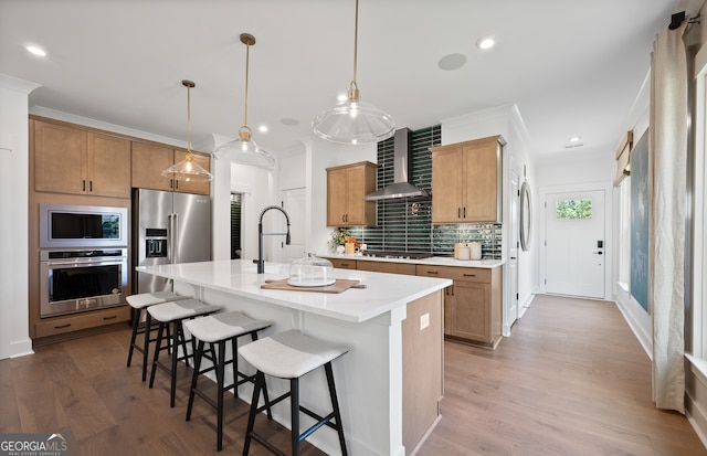 kitchen with stainless steel appliances, wall chimney range hood, pendant lighting, light hardwood / wood-style flooring, and an island with sink