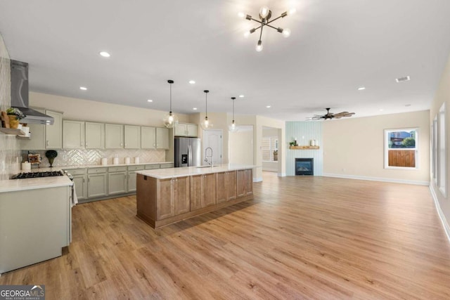 kitchen featuring stainless steel appliances, wall chimney range hood, light hardwood / wood-style flooring, pendant lighting, and a center island with sink