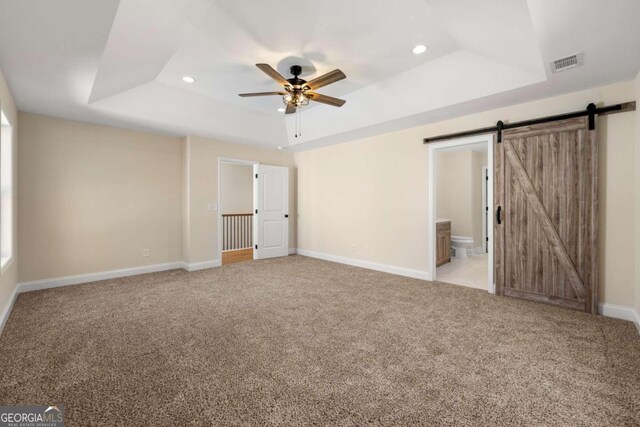 unfurnished bedroom featuring ensuite bath, a tray ceiling, light colored carpet, ceiling fan, and a barn door