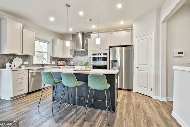 kitchen with light wood-type flooring, wall chimney exhaust hood, stainless steel appliances, a kitchen island, and hanging light fixtures