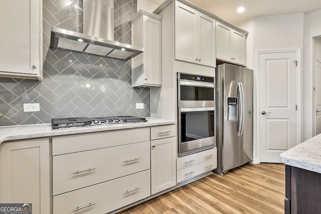 kitchen with backsplash, wall chimney exhaust hood, appliances with stainless steel finishes, light hardwood / wood-style floors, and light stone counters