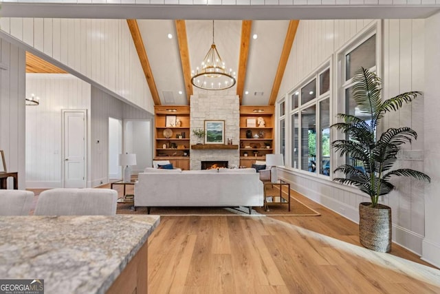 living room featuring a notable chandelier, lofted ceiling with beams, wood-type flooring, a stone fireplace, and built in shelves