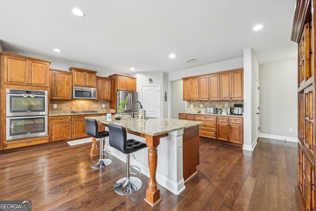 kitchen featuring a kitchen island with sink, dark wood-type flooring, a kitchen breakfast bar, light stone countertops, and appliances with stainless steel finishes