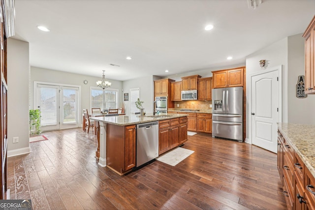 kitchen with dark wood-type flooring, hanging light fixtures, stainless steel appliances, a notable chandelier, and a kitchen island with sink