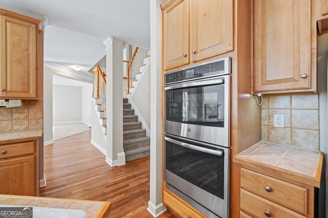 kitchen featuring tile counters, double oven, light wood-type flooring, decorative backsplash, and ornamental molding