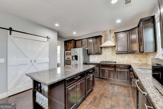 kitchen with wall chimney exhaust hood, a barn door, dark hardwood / wood-style floors, appliances with stainless steel finishes, and a kitchen island