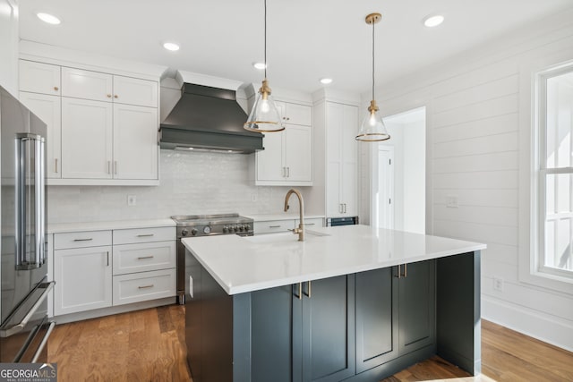 kitchen with sink, appliances with stainless steel finishes, a kitchen island with sink, decorative light fixtures, and custom exhaust hood