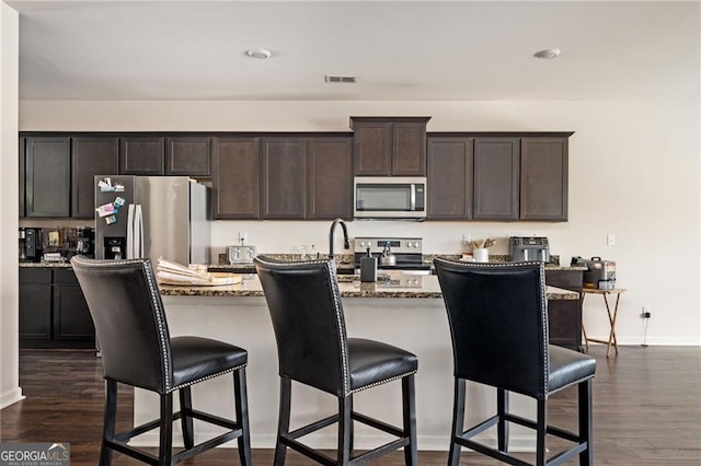 kitchen featuring a kitchen breakfast bar, an island with sink, dark hardwood / wood-style flooring, light stone counters, and stainless steel appliances