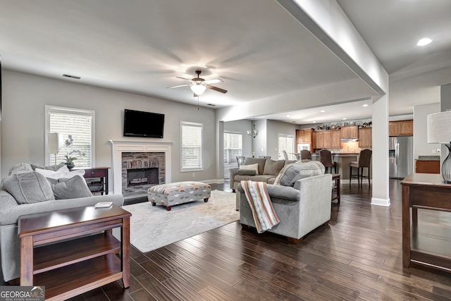 living room with a fireplace, ceiling fan with notable chandelier, a wealth of natural light, and dark wood-type flooring