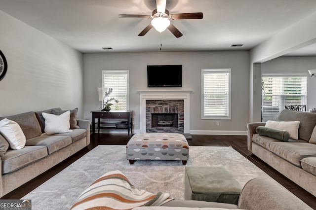 living room with ceiling fan, a healthy amount of sunlight, dark hardwood / wood-style flooring, and a stone fireplace