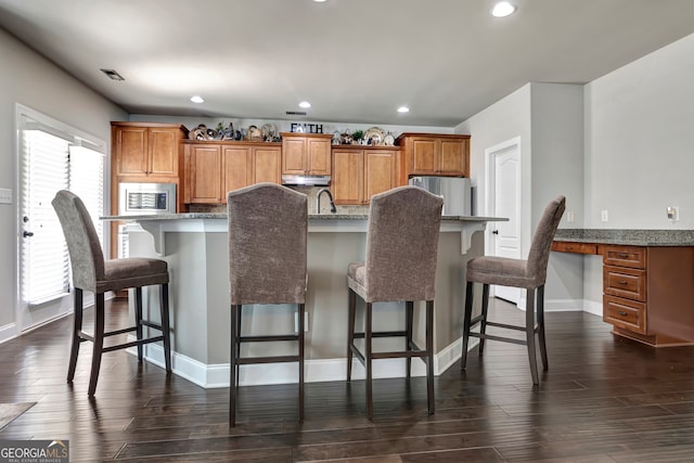 kitchen with dark wood-type flooring, an island with sink, a kitchen bar, decorative backsplash, and appliances with stainless steel finishes