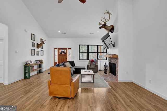 living room with ceiling fan, a brick fireplace, a towering ceiling, and light hardwood / wood-style floors