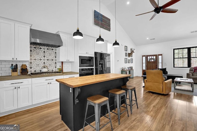 kitchen with a kitchen bar, butcher block countertops, white cabinetry, black appliances, and wall chimney range hood