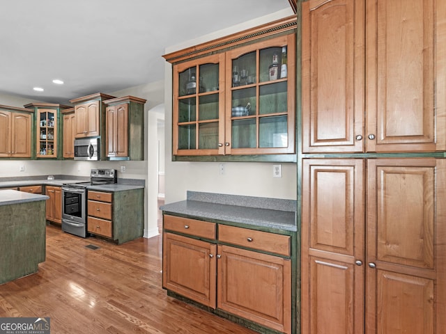 kitchen featuring recessed lighting, stainless steel appliances, wood finished floors, dark countertops, and glass insert cabinets