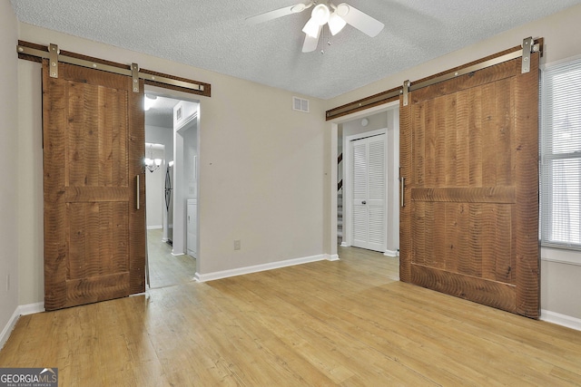 unfurnished bedroom with a textured ceiling, ceiling fan, a barn door, and light hardwood / wood-style flooring