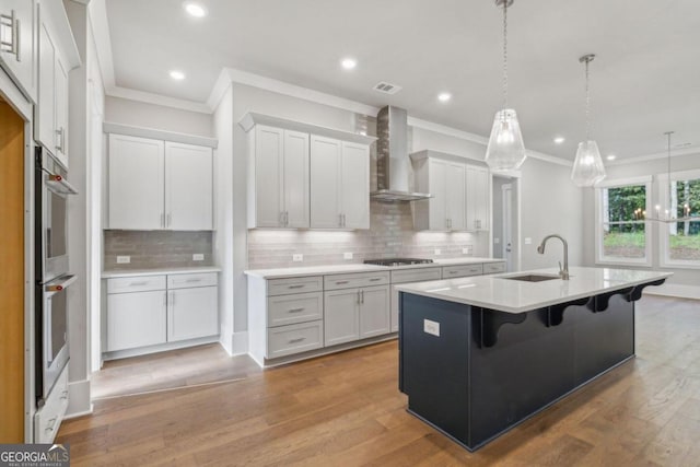 kitchen with hardwood / wood-style floors, white cabinets, wall chimney range hood, sink, and an island with sink